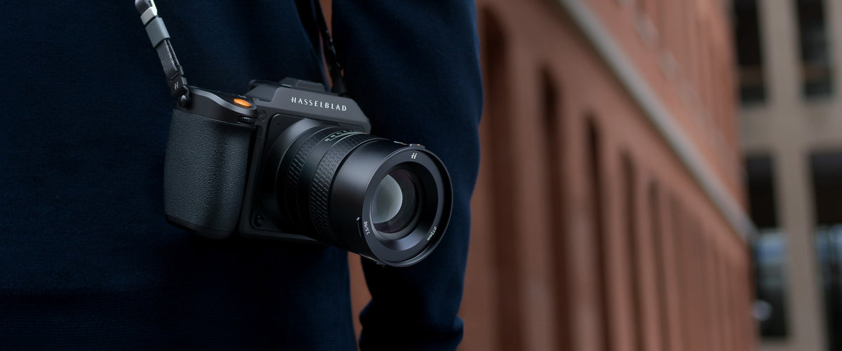 Hasselblad-Kamera mit Objektiv hängt am Schultergurt einer Person vor moderner Architektur