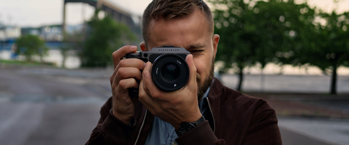Mann fotografiert im Freien mit einer Hasselblad-Kamera vor unscharfem Stadt- und Baumhintergrund