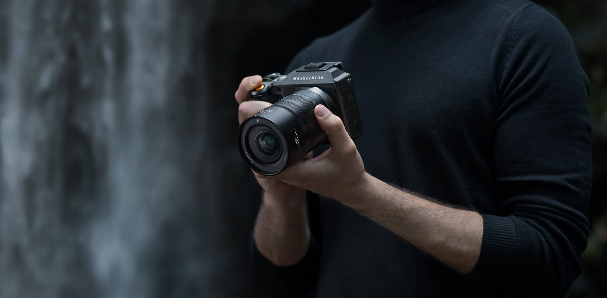 Person in schwarzem Pullover hält eine Hasselblad-Kamera mit Objektiv vor dunklem Hintergrund